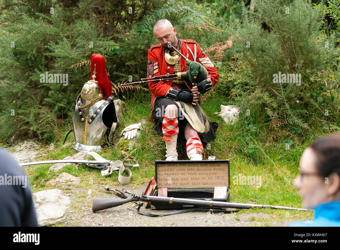 Man busking dressed in Scottish Soldier Outfit sitting by his armor and playing bagpipes at the entrance to the monastic site in Glendalough Valley Stock Photo