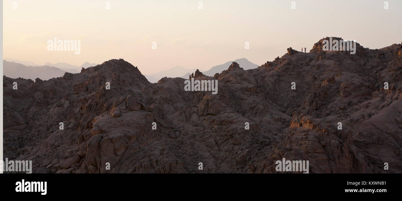 two people in the distance standing on top of a mountain range in evening light having a photograph taken. Stock Photo