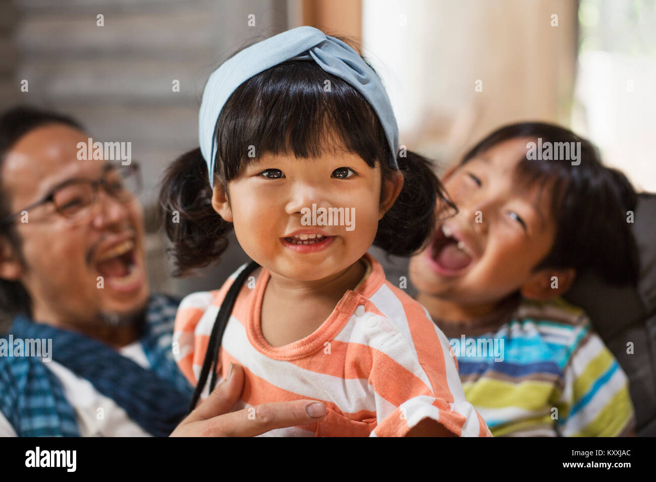 Man, boy and young girl with black pigtails wearing blue hairband sitting on a grey sofa, laughing. Stock Photo