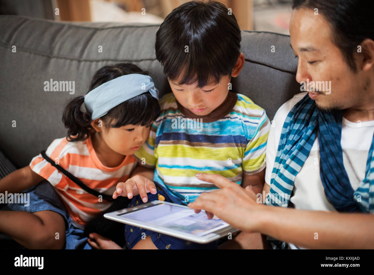 Man, boy and young girl sitting on a grey sofa, looking at digital tablet. Stock Photo