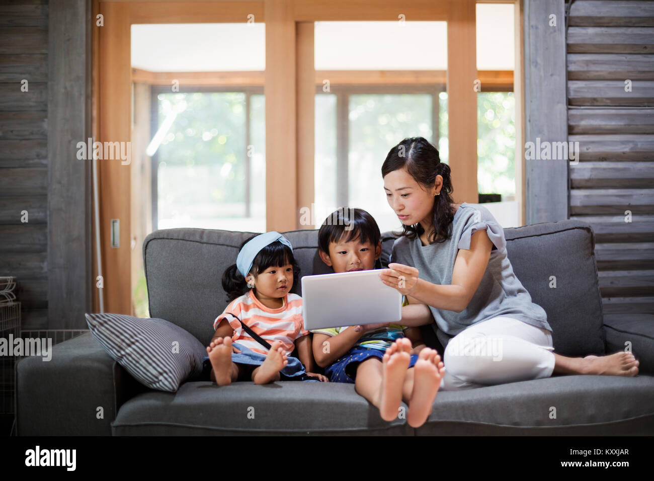 Woman, boy and young girl sitting on a grey sofa, looking at digital tablet. Stock Photo