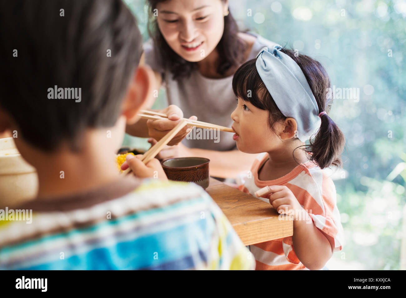 Young girl with black pigtails and blue hairband, boy and woman sitting at a table. Stock Photo