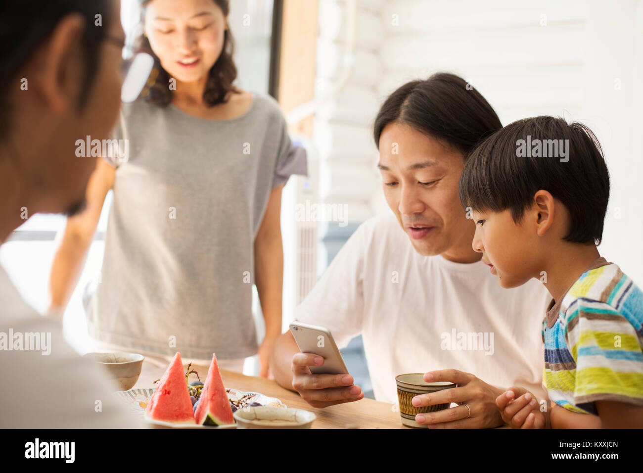 Two men, boy and woman indoors round a table, man and boy looking at mobile phone. Stock Photo