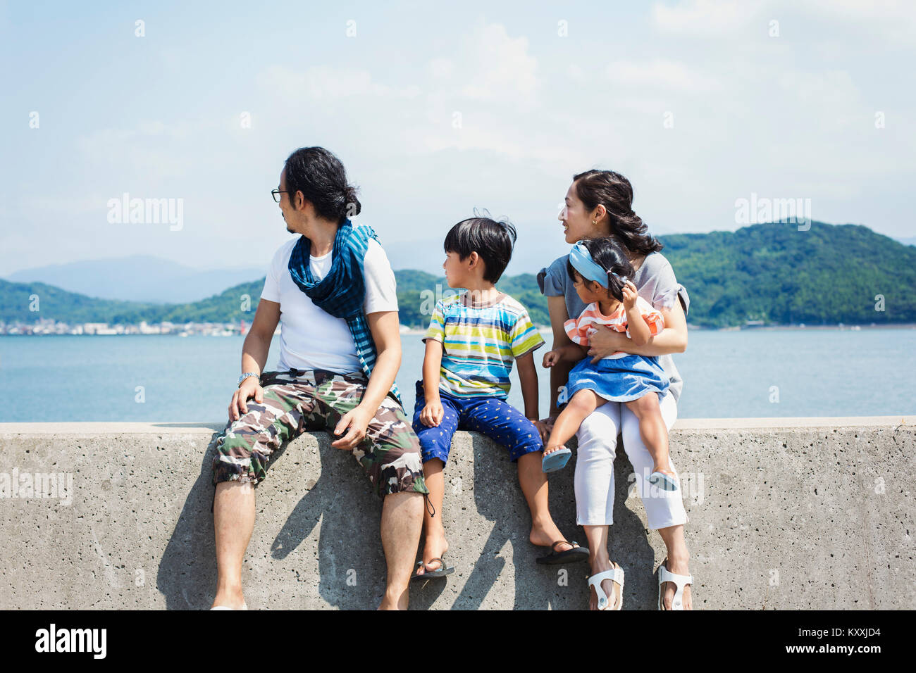 Family, man, boy and woman with young girl on her lap sitting side by side on a wall by the ocean. Stock Photo