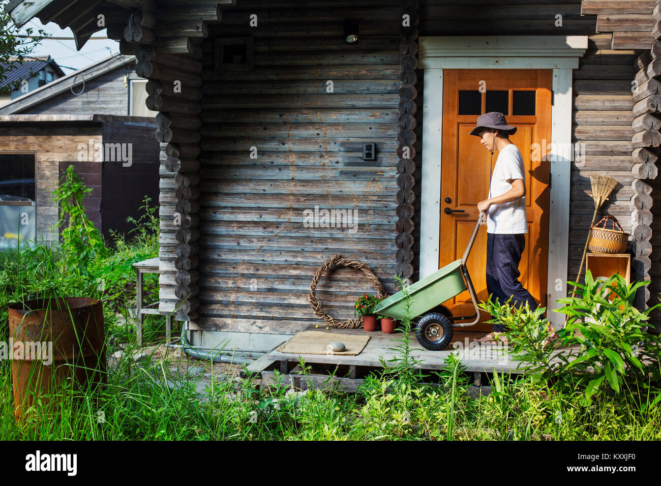 Man wearing hat standing outside wooden garden shed, pushing green wheelbarrow. Stock Photo