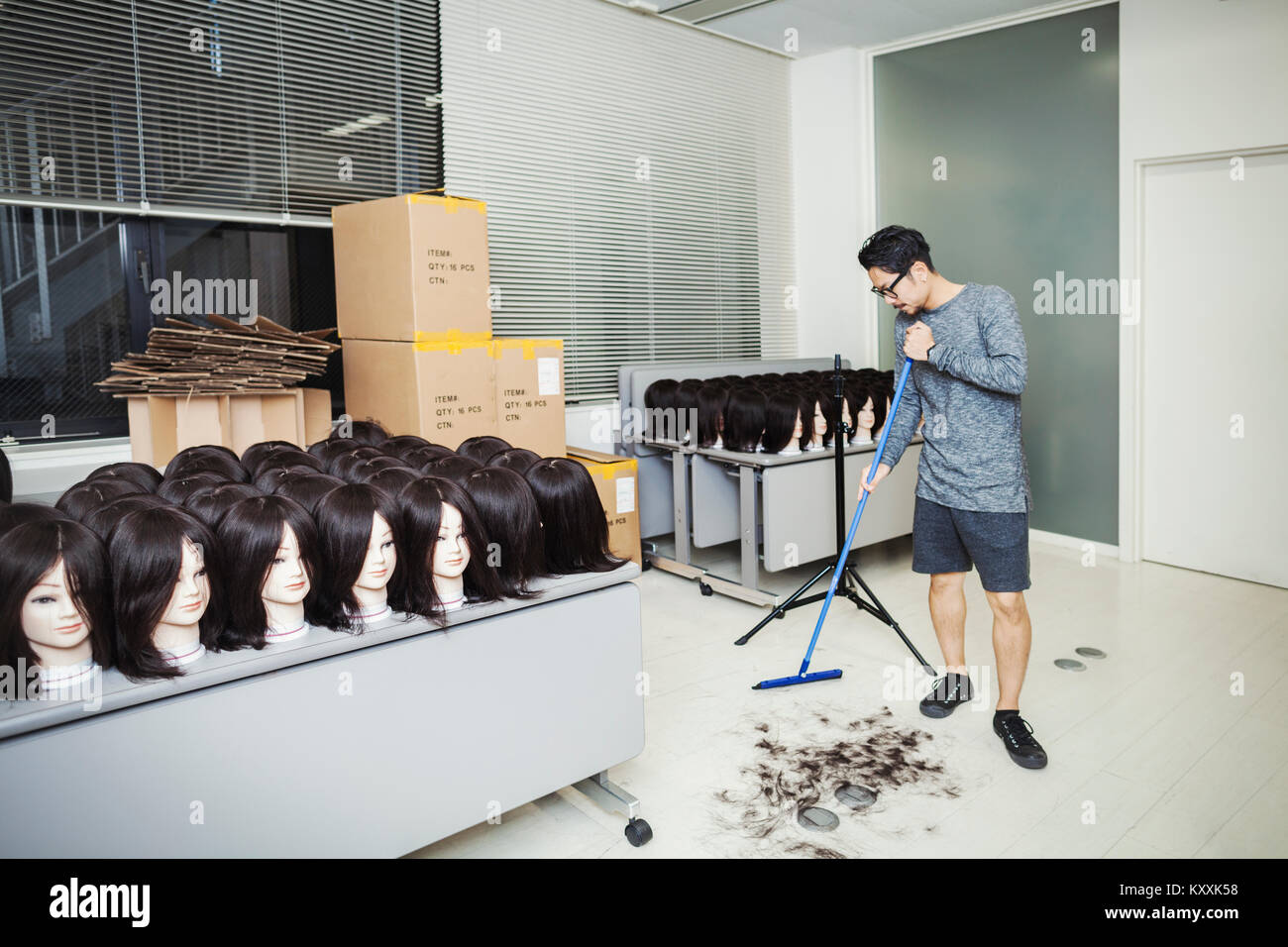 Bearded man wearing glasses standing indoors, sweeping hair on floor, large group of mannequin heads with brown wigs on tables. Stock Photo