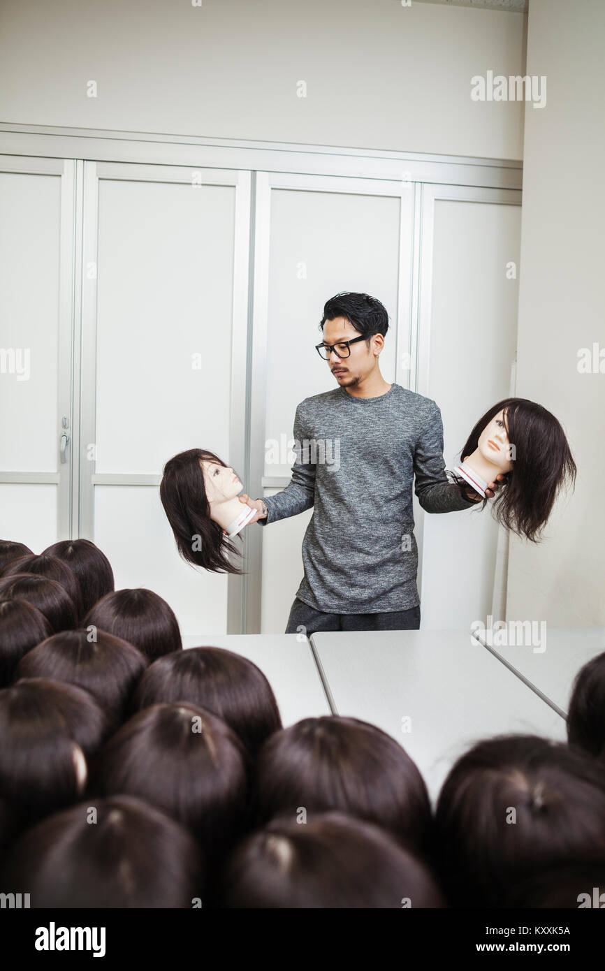 Bearded man wearing glasses standing indoors, holding mannequin head with brown wig in each hand. Stock Photo