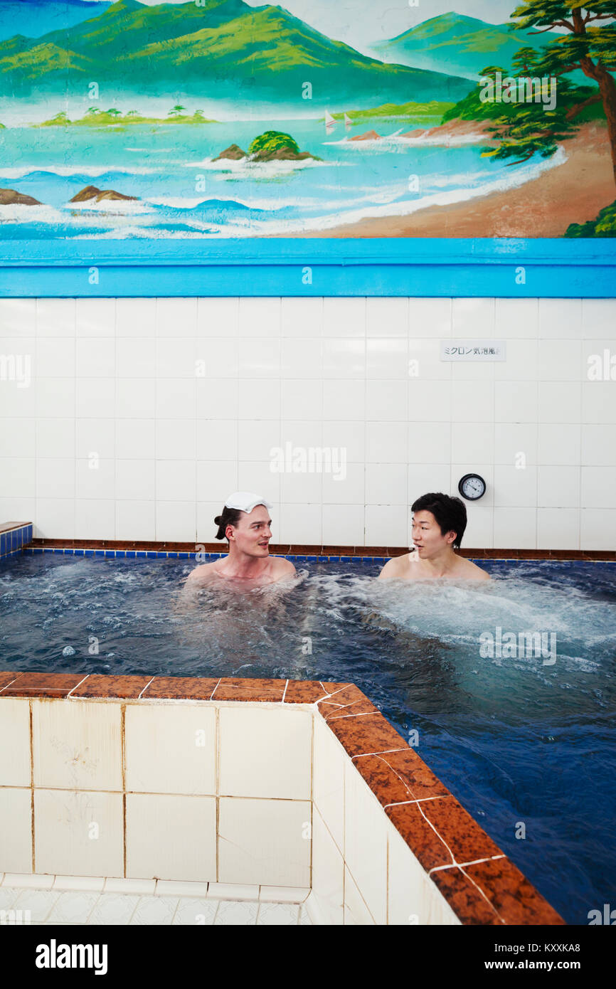 A young Caucasian man and Japanese man seated in a deep pool of moving water in a public bath house. Stock Photo