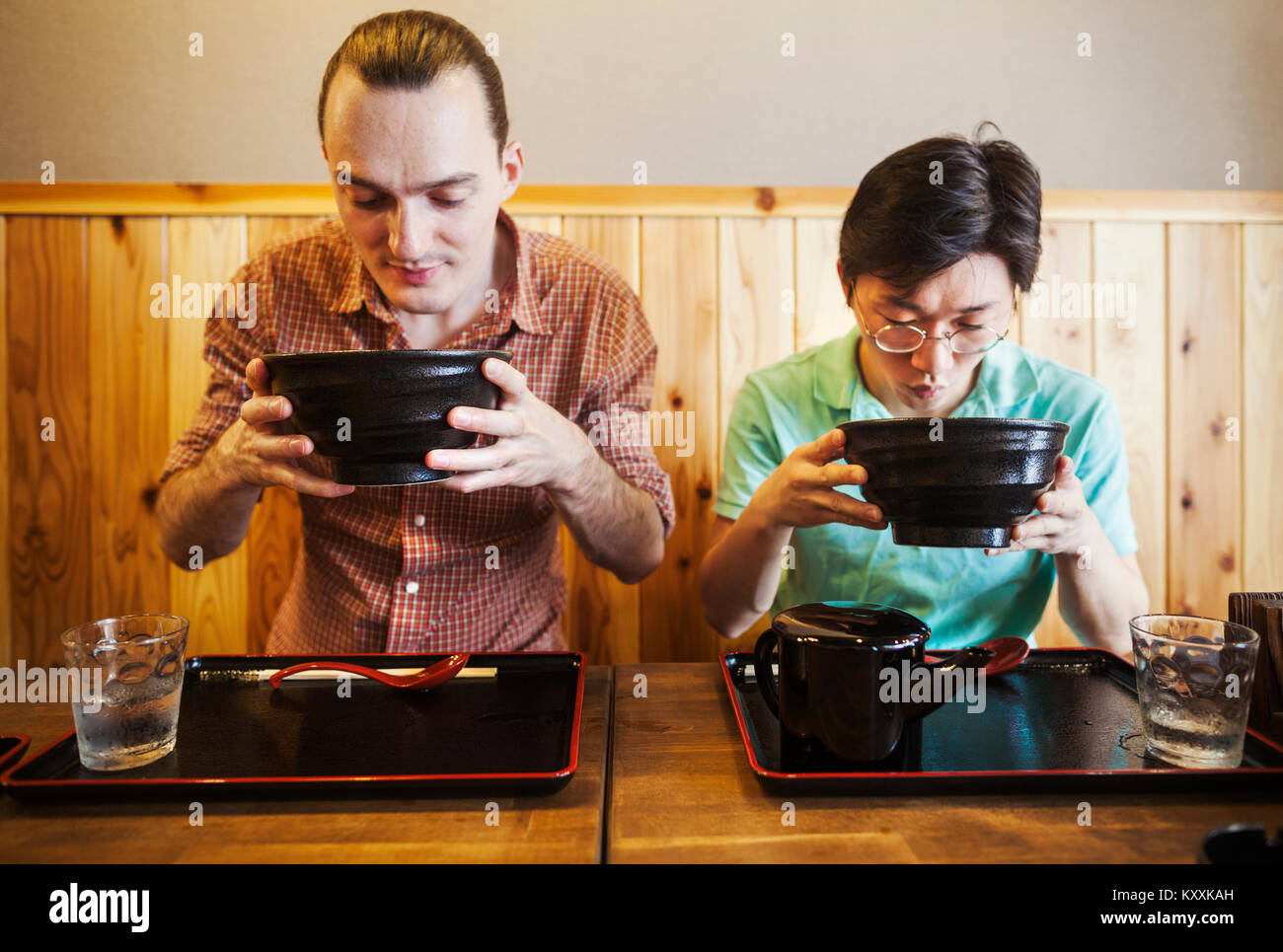 Two people in a noodle cafe lifting bowls of soba noodles. A western man and a Japanese man. Stock Photo