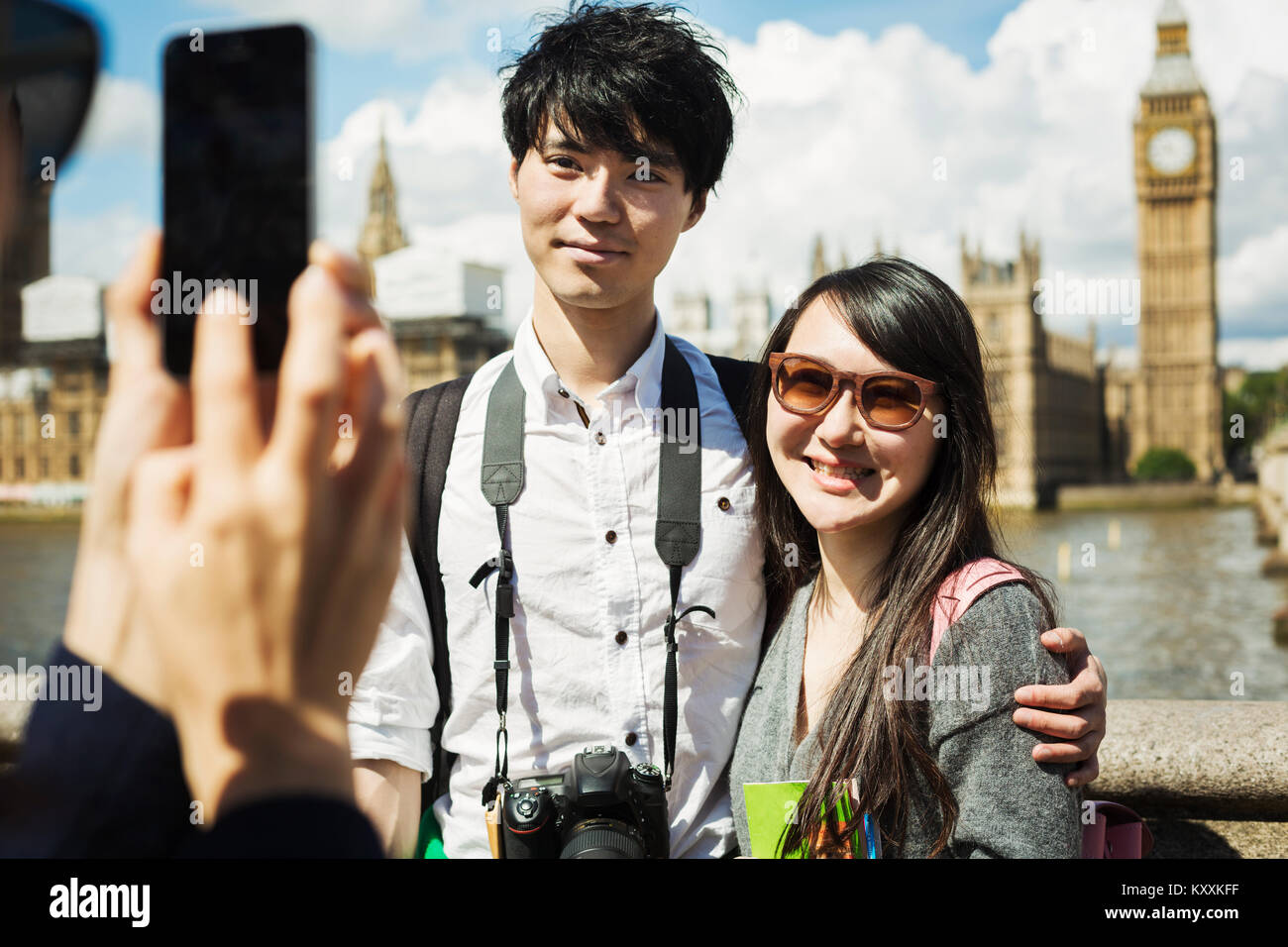 Smiling woman with black hair taking picture of couple with smartphone, standing on Westminster Bridge over the River Thames, London, with the Houses  Stock Photo