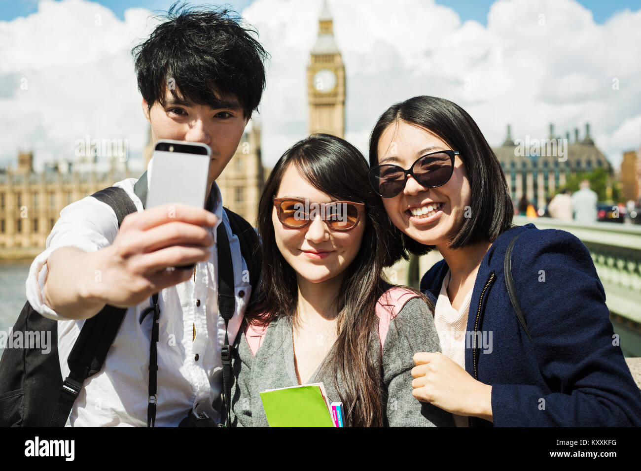 Smiling man and two women with black hair taking selfie with smartphone, standing on Westminster Bridge over the River Thames, London, with the Houses Stock Photo