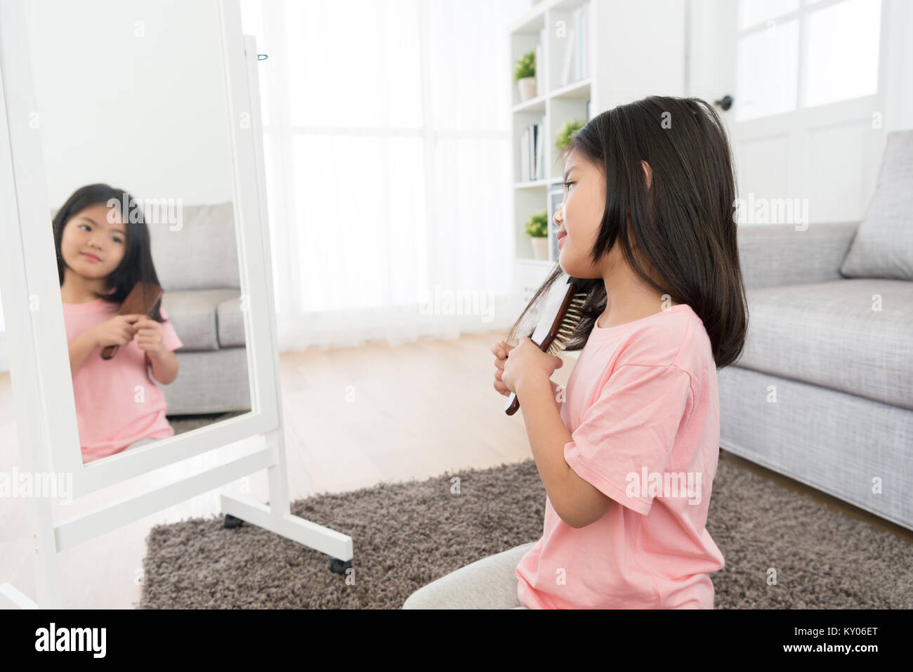 attractive young female kid child sitting on floor looking at mirror and using brush combing hair by herself in living room. Stock Photo