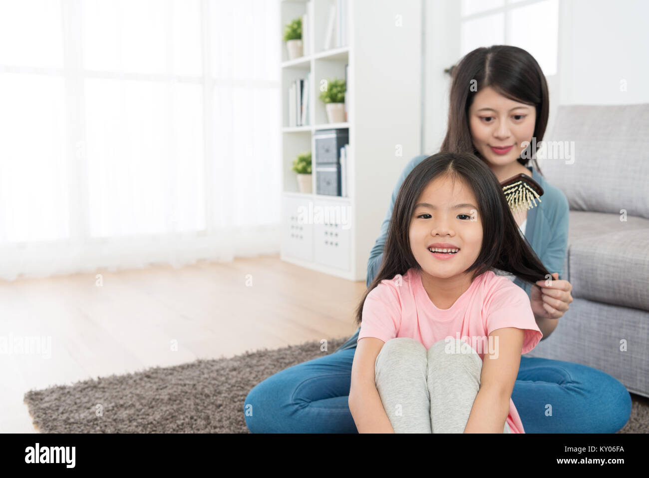 beautiful smiling children girl sitting on floor looking at camera when mother using brush combing her hair at home in morning. Stock Photo