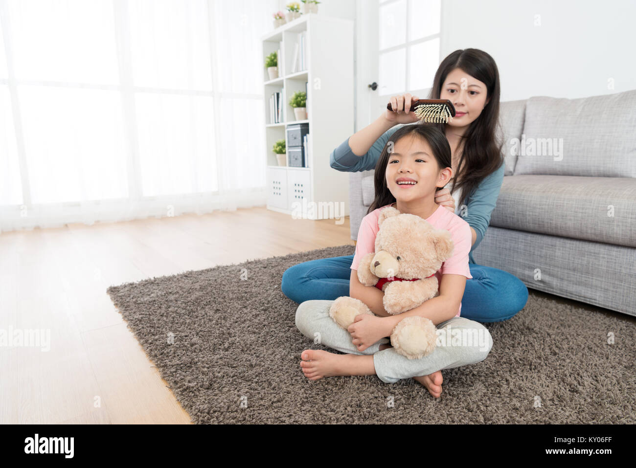 happy cute girl children holding teddy bear sitting on living room floor and pretty young mother combing her hair in morning. Stock Photo