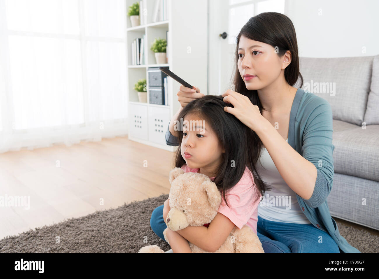 young cute little girl holding teddy toy sitting on floor in morning and face play grimace looking at mirror when mother combing her hair. Stock Photo