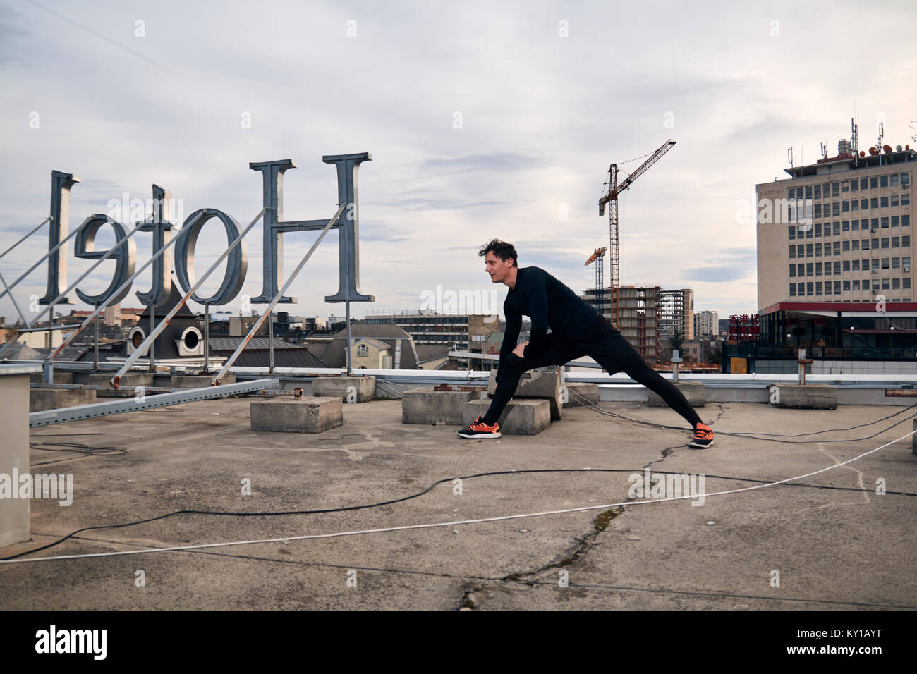 one young man workout stretching outdoors on rooftop. buildings crane behind, urban area city cityscape. Hotel sign backwards. Stock Photo
