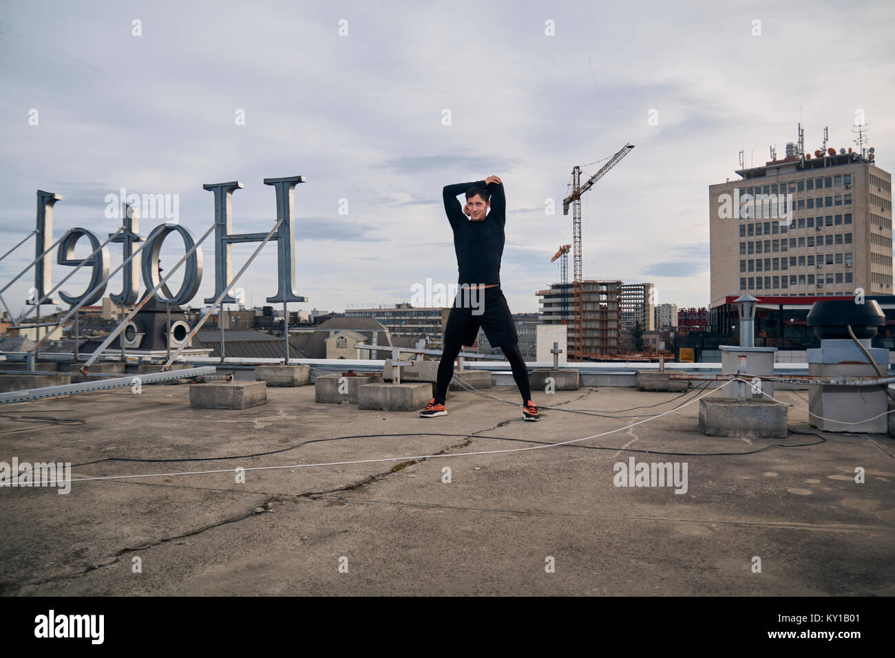 one young man stretching outdoors on rooftop. Buildings crane behind, cityscape, urban area. 'Hotel' sign spelled backwards. Stock Photo
