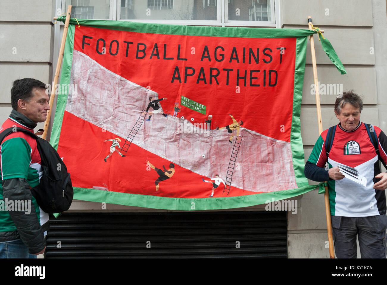 Two protestors wearing Palestine solidarity colours with large banner 'Football against Apartheid'showing footballers breaching Israel/ Palestine wall Stock Photo