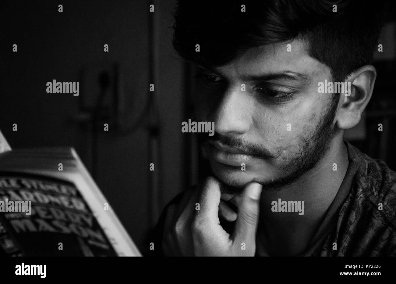 Black and white close-up portrait of a young adult Indian boy reading a book with hand on chin, thinking gesture. Stock Photo