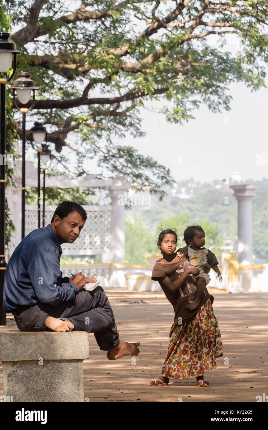 Indifference the root of Inequality: Gentleman sitting on bench looks away from a poor, shabbily dressed young girl holding her sibling. Stock Photo