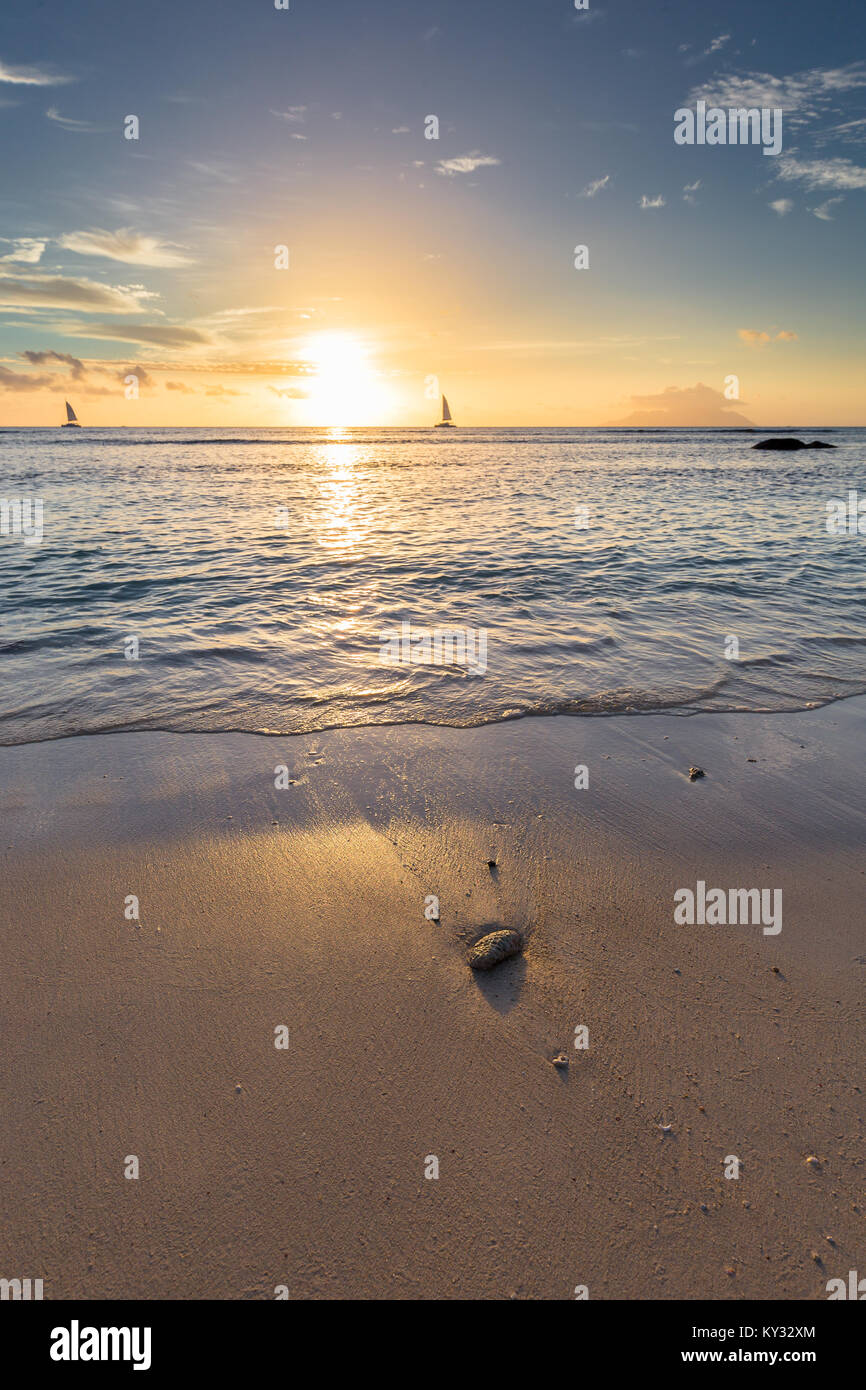 Sunset over the ocean with two sailboats on the horizon and a piece of coral on the beach Stock Photo