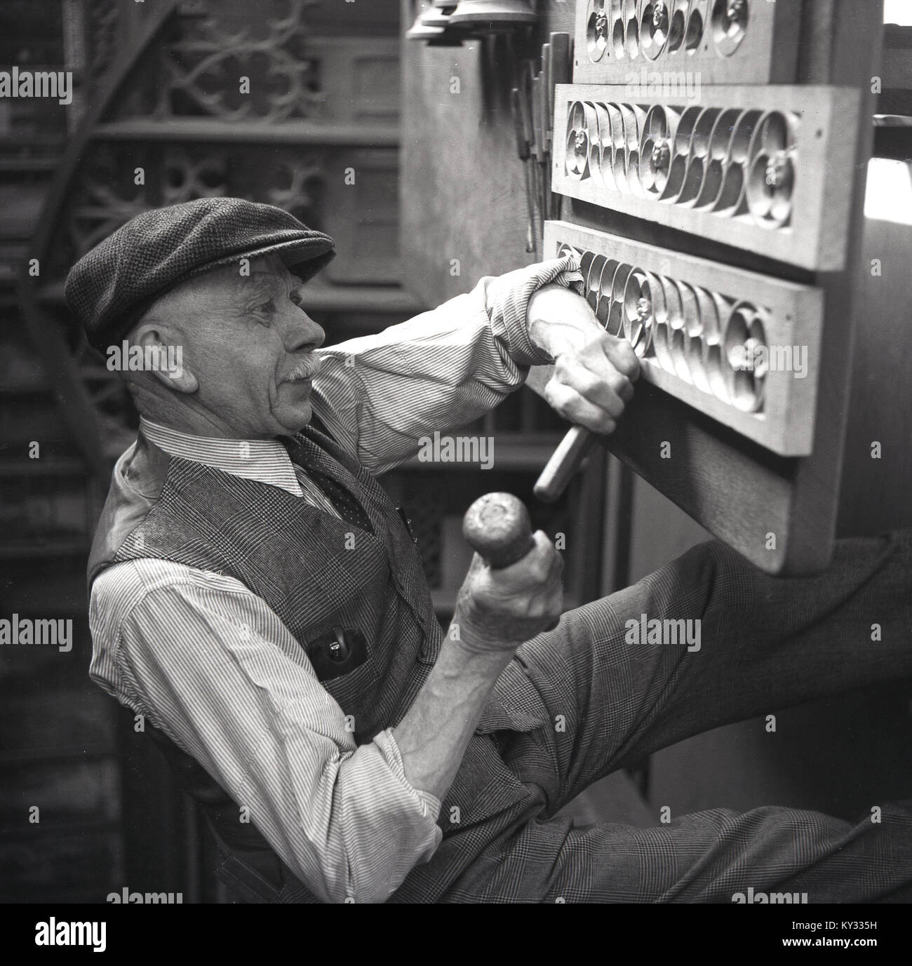 1950s, historical, artisian woodworking...an elderly man wearing a waistcoat, suit trousers and a flat clap using a mallet and chisel to carve out shapes on wooden blocks fixed above him, England, UK. Stock Photo