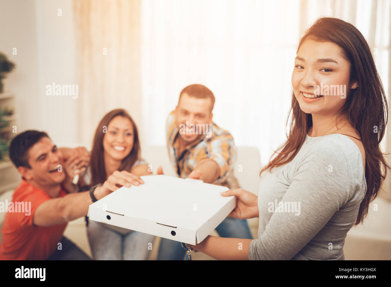 Four cheerful friends enjoying pizza together at home party. Selective focus. Focus on foreground, on Japanese girl. She is holding box with pizza and Stock Photo