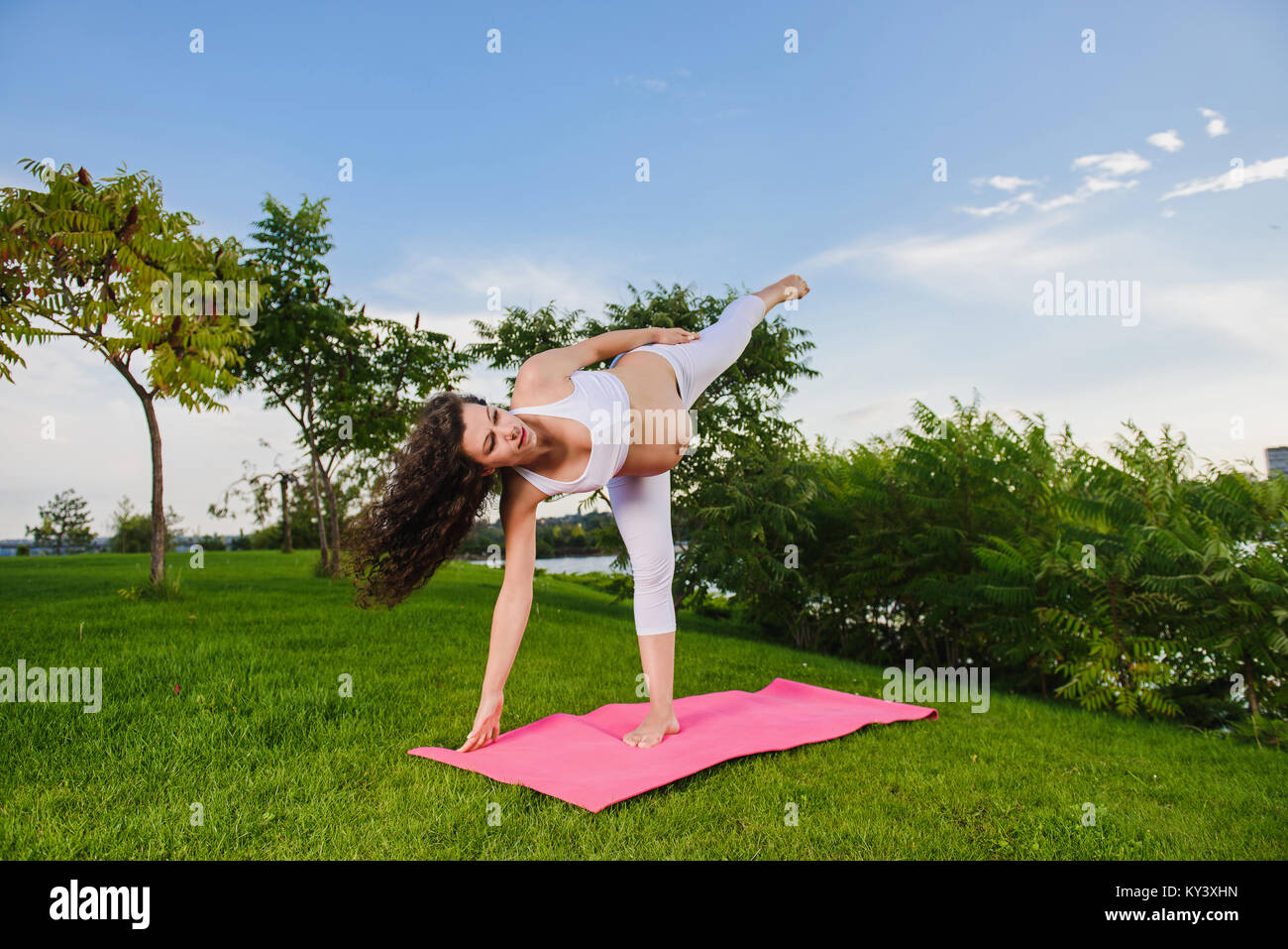 Pregnant woman doing prenatal yoga on nature. Ardha candrasana, pose of the half moon. Woman in park on grass. Stock Photo