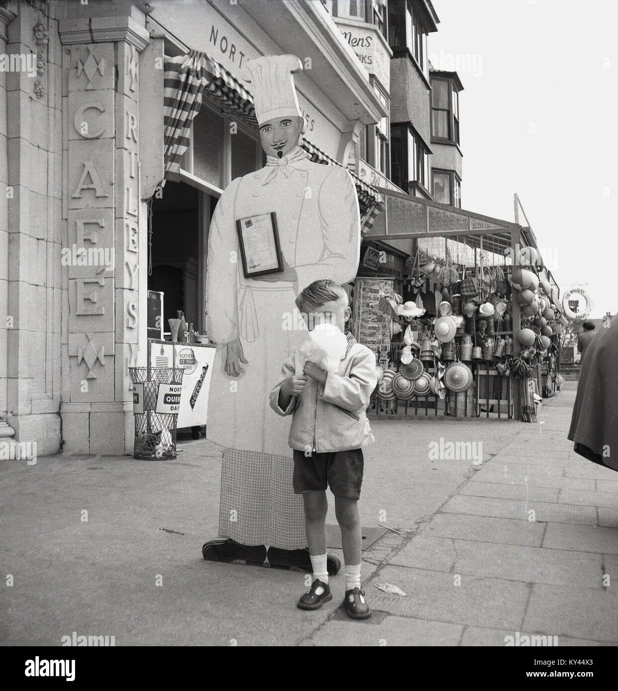 1950s, historical picture, a young boy in short trousers enjoys his seaside treat of candy floss, with a large promotional cut-out of a chef with hat standing behind with him. Stock Photo