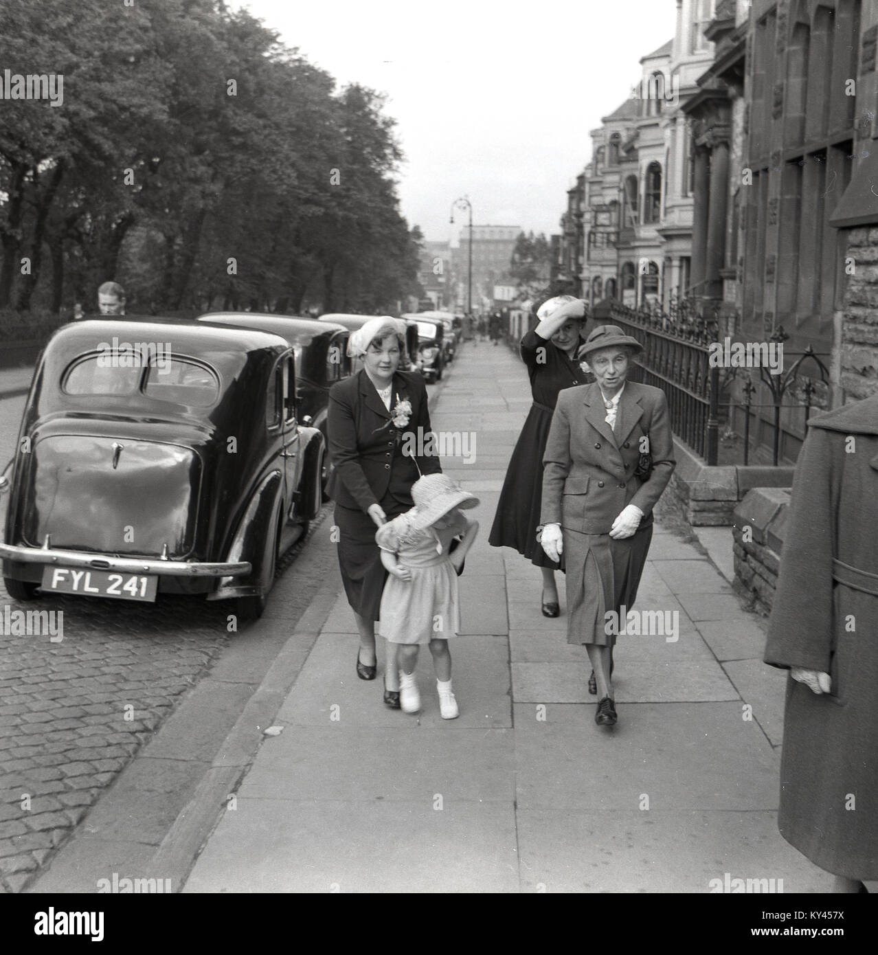 1950s, historical, three well-dressed ladies and a child in dress and bonnet walk up a pavement past the waiting cars towards a church to attend a wedding, England, UK. Stock Photo