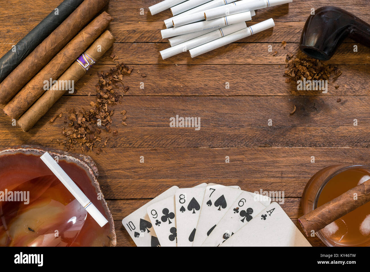 Smoking and tobacco products, cigars, cigarettes and a pipe with tobacco, on top of a wooden background, with playing cards, ashtray and drink Stock Photo