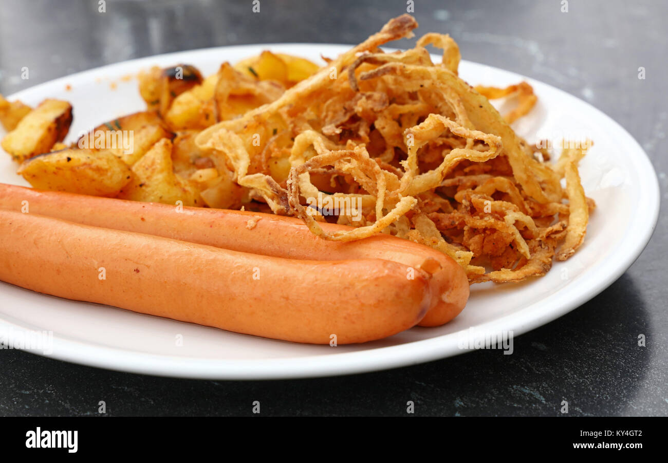 Close up portion of two boiled sausages with roasted potato and fried onion rings on white plate over grey table, low angle view Stock Photo