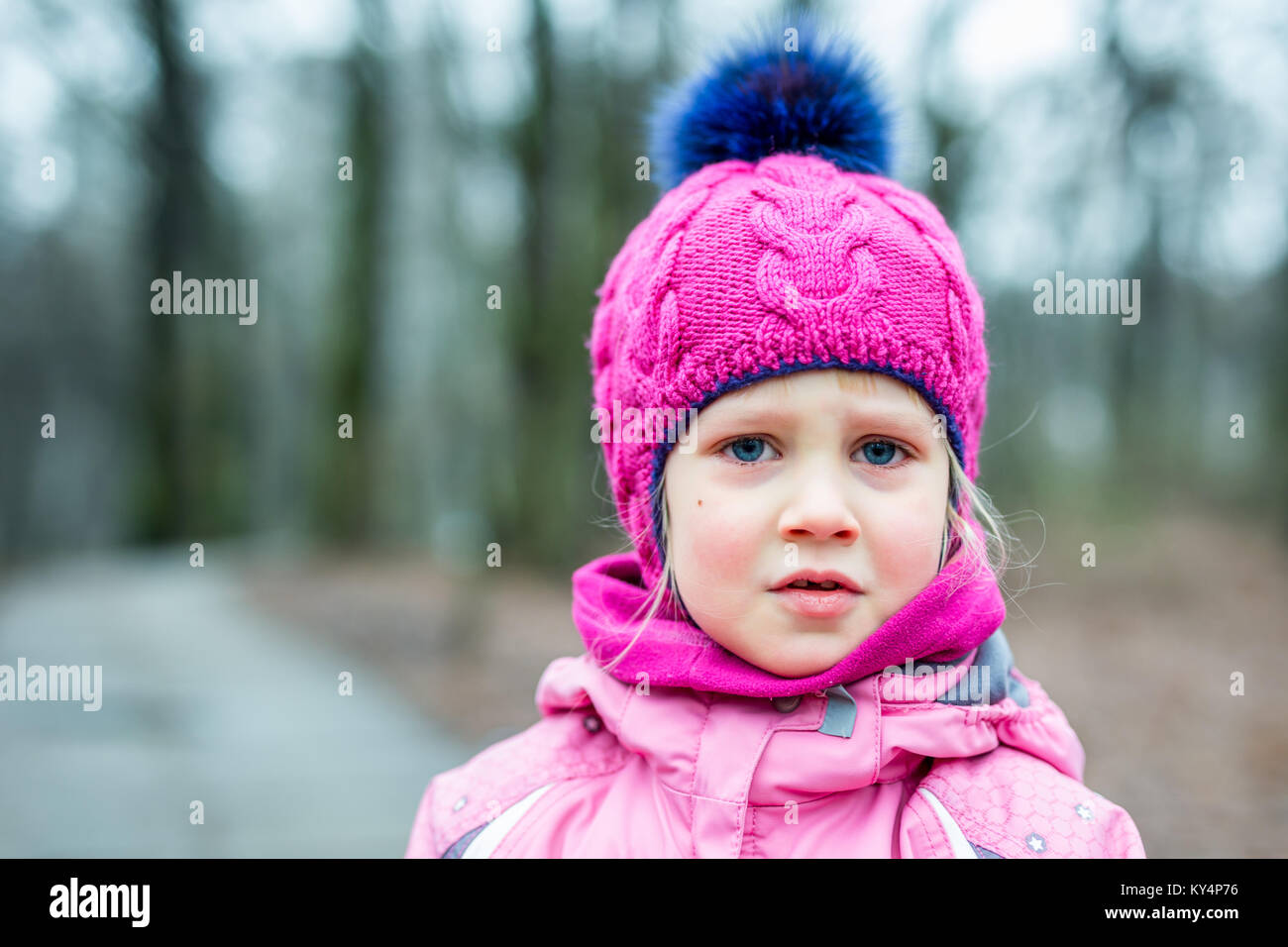 Portrait of little cute caucasian girl in pink knitted hat crying , being upset or lost in a forest or city park . Stock Photo