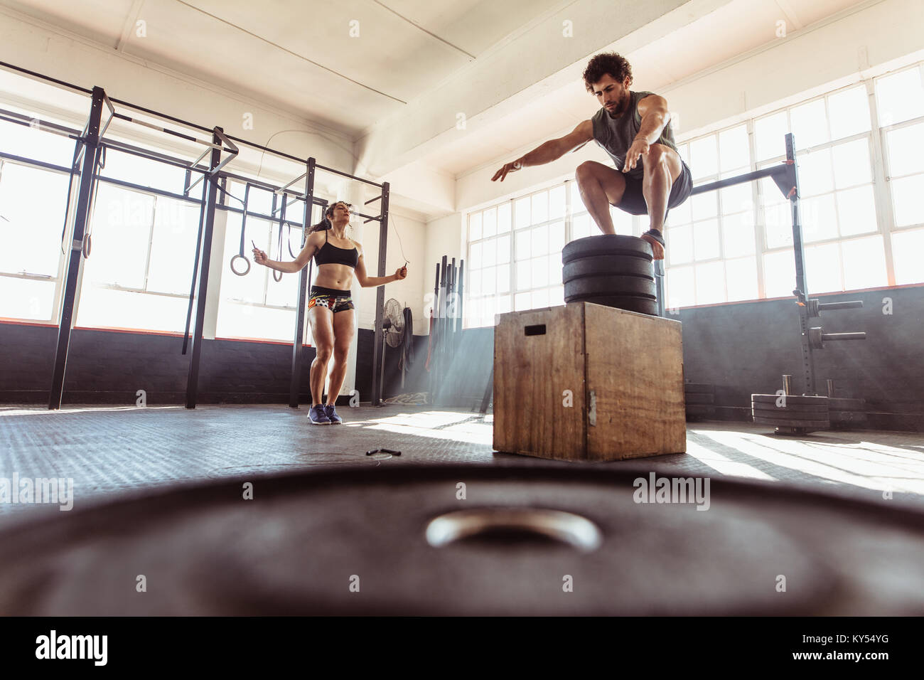 Fit young man box jumping with woman exercising with skipping ropes at a cross training style gym. Couple during intense workout session at health clu Stock Photo
