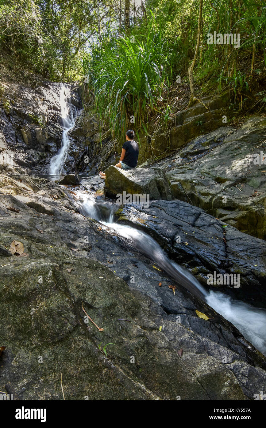 Young woman sat on rocks looking at a jungle waterfall contemplating ...