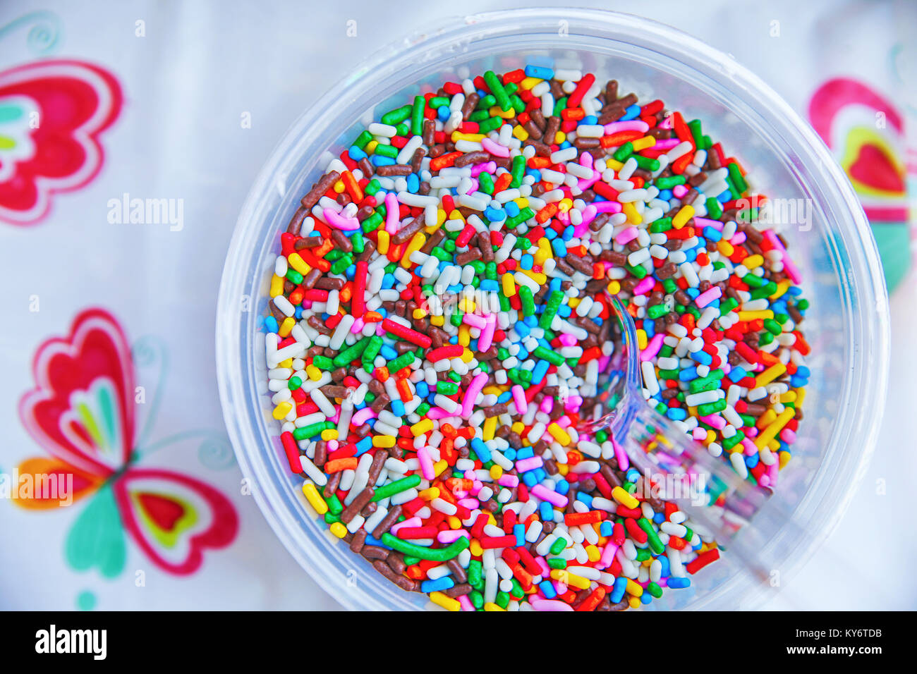 a bowl full of colorful candy sprinkles with a clear plastic spoon in them and a plastic tablecloth Stock Photo