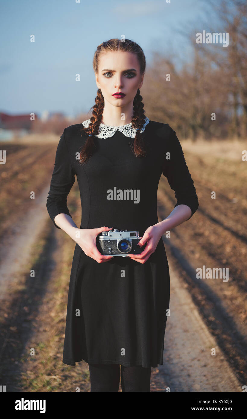 Outdoor portrait of a cute young girl in old-fashioned black dress with vintage film camera in hands Stock Photo