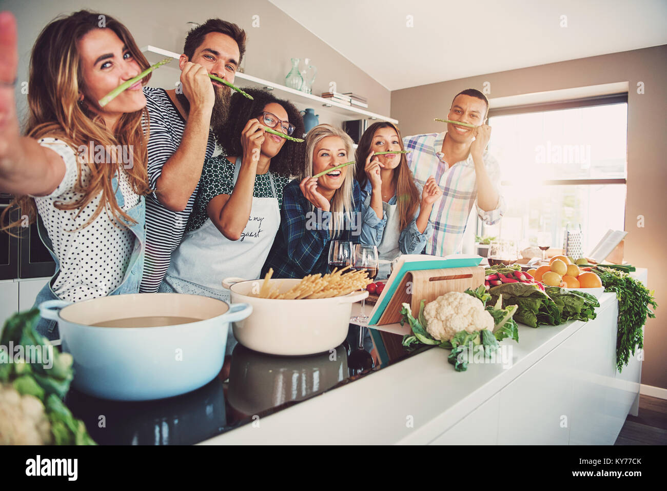 Friends wearing asparagus stalks under their noses while standing in front of table full of pots, pasta noodles and vegetables Stock Photo