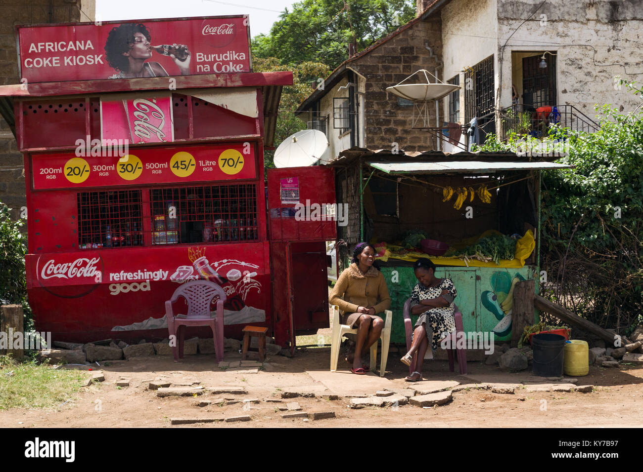 Two Kenyan women sit talking outside metal stalls selling Coca Cola and fruit and vegetables, Nairobi, Kenya, East Africa Stock Photo