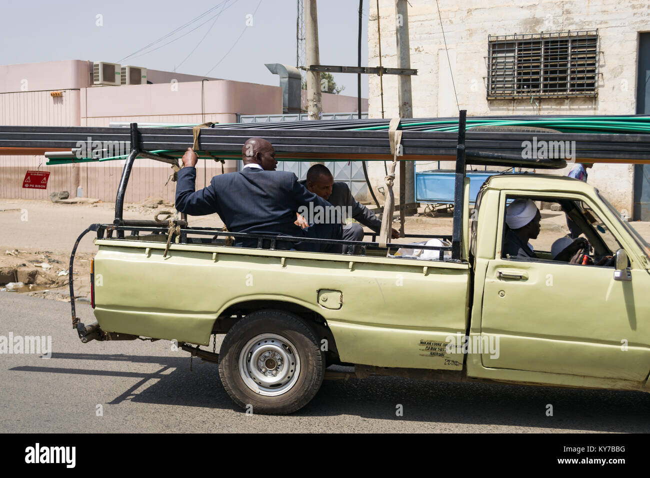 Two Kenyan men in suits sit in the back of a pickup truck as it transports steel framework on a road, Nairobi, Kenya, East Africa Stock Photo