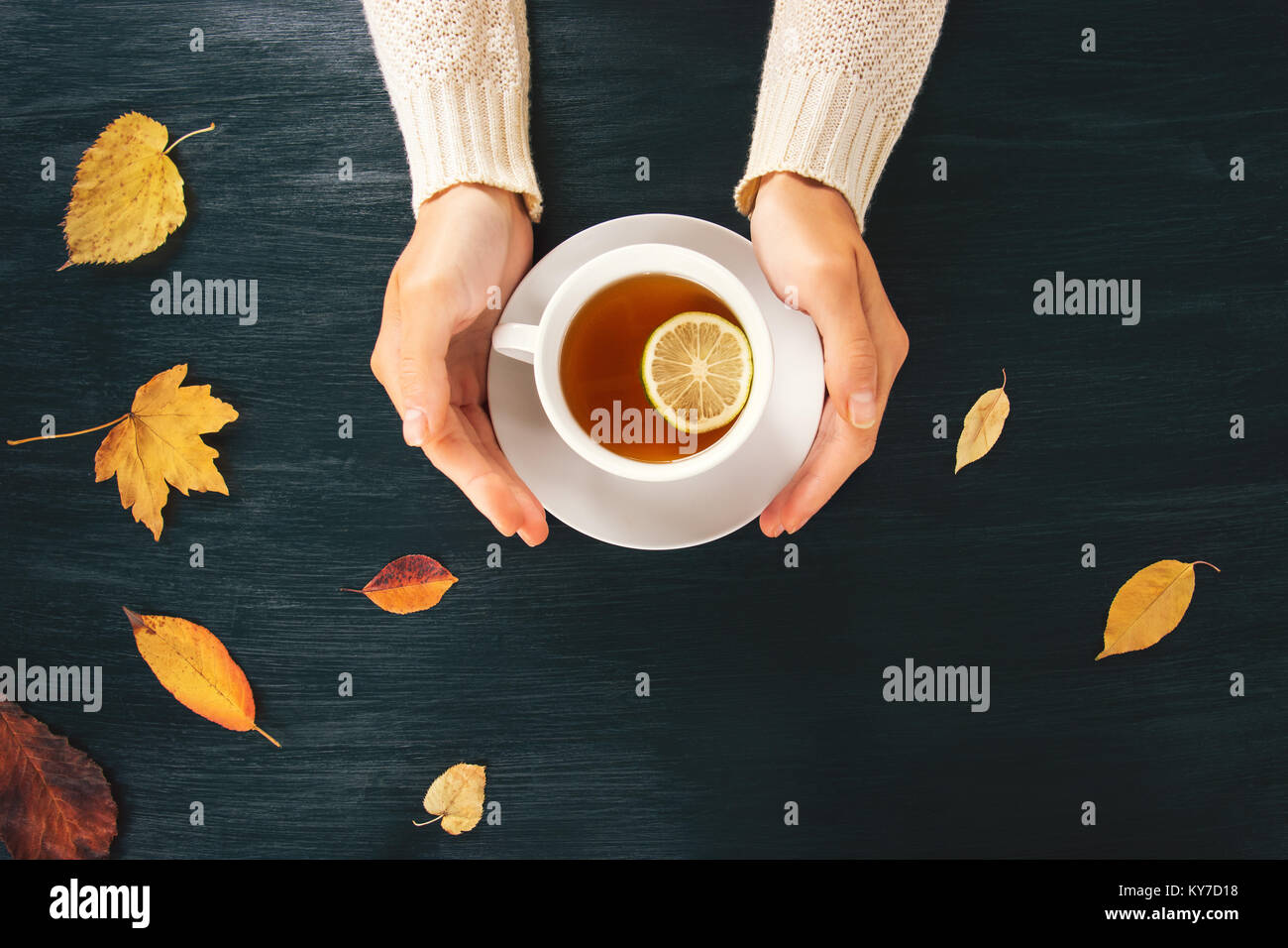 Woman hands holding hot tea lemon cup on wooden table with autumn leaves seasonal concept top view trendy style Stock Photo