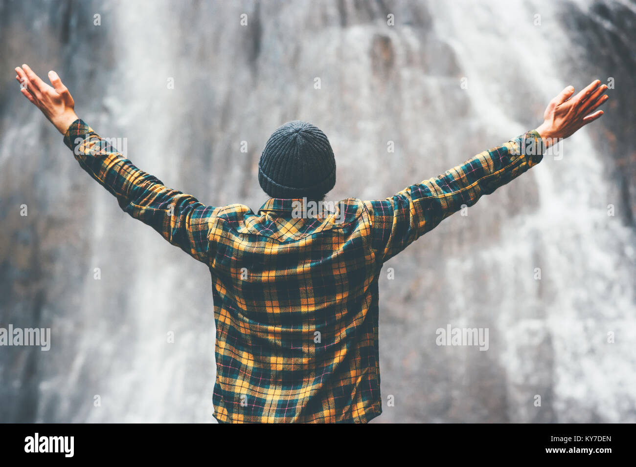 Man enjoying waterfall raised hands Travel Lifestyle adventure concept vacations into the wild wearing cozy shirt and hat Stock Photo