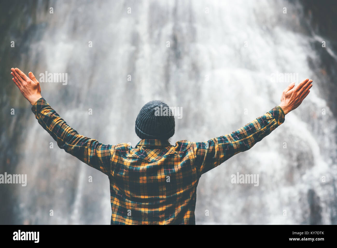 Man enjoying waterfall raised hands Travel Lifestyle and success concept vacations into the wild wearing cozy shirt and hat Stock Photo