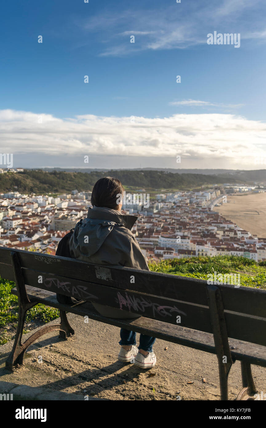 woman sitting on a bench enjoying the view of the coast and the town of Nazare in Portugal. Stock Photo