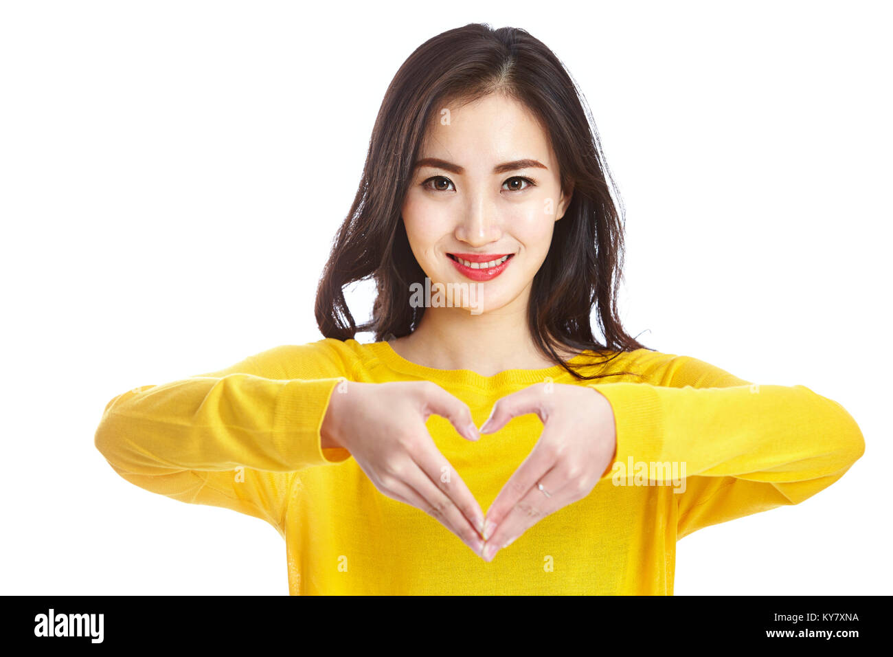 studio shot of a young and beautiful asian woman forming a heart shape with hands, looking at camera smiing, isolated on white background. Stock Photo