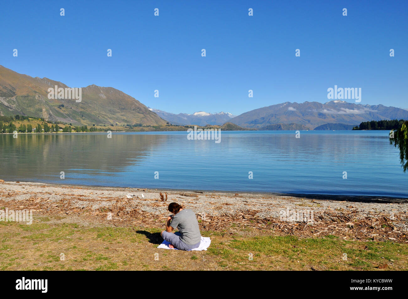 Female reading by Lake Wanaka with mountains beyond on blue sky sunny day. Otago region, South Island, New Zealand Stock Photo