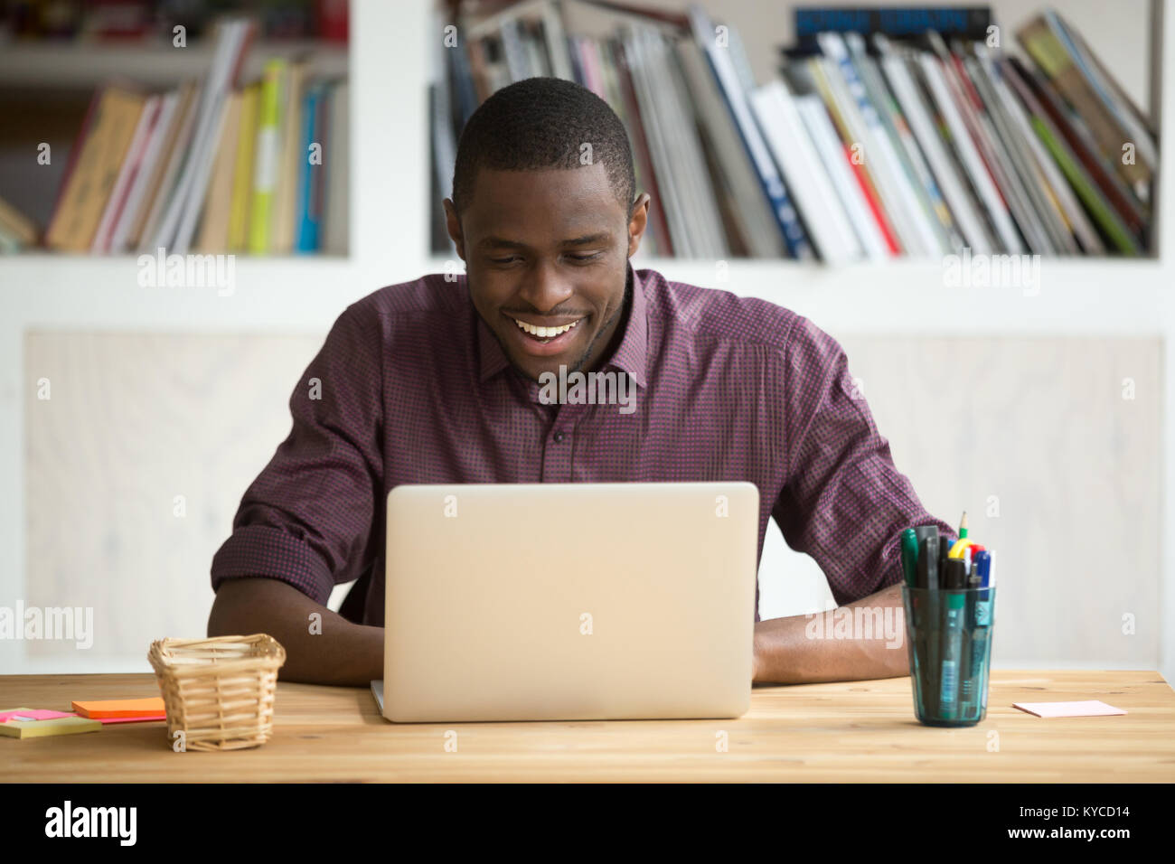 Smiling african-american man using laptop sitting at home office desk, happy excited black e business owner looking at computer screen pleased by read Stock Photo