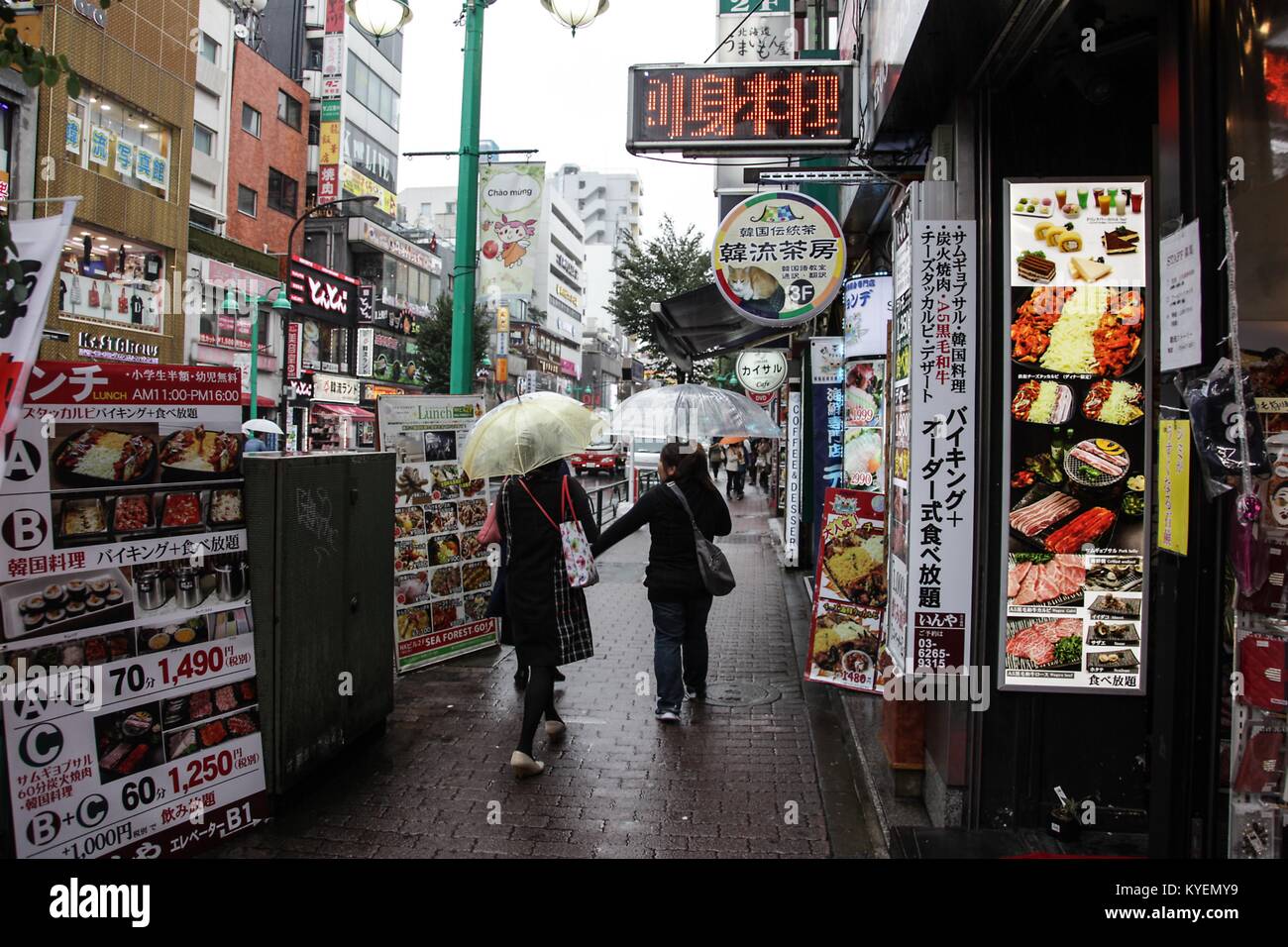 People carrying umbrellas walk past signs for street food in the Kabukicho Red Light District, Shinjuku, Tokyo, Japan on a rainy day, October 16, 2017. () Stock Photo