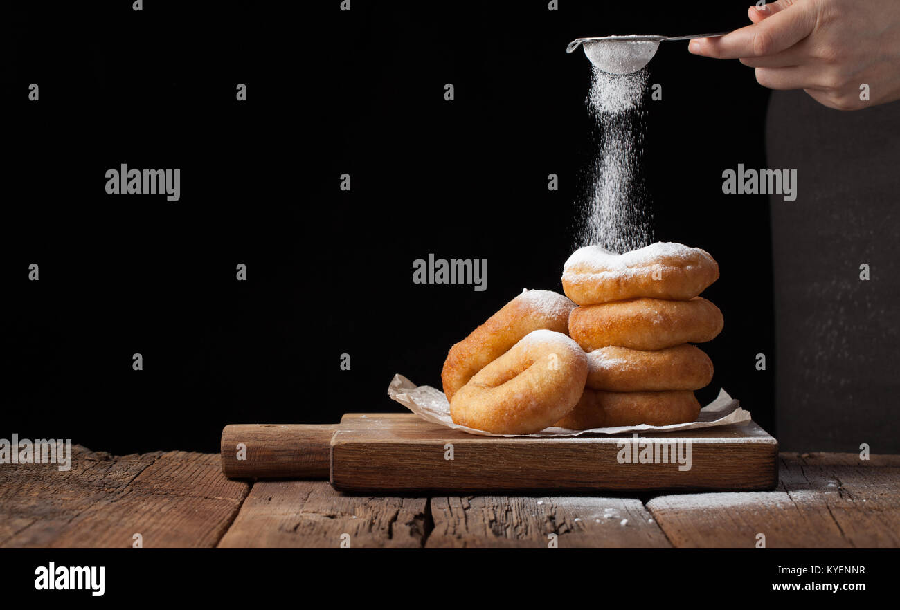 Baker sprinkles sweet donuts with powder sugar on black background. Delicious, but unhealthy food on the old wooden table with copy space. Stock Photo