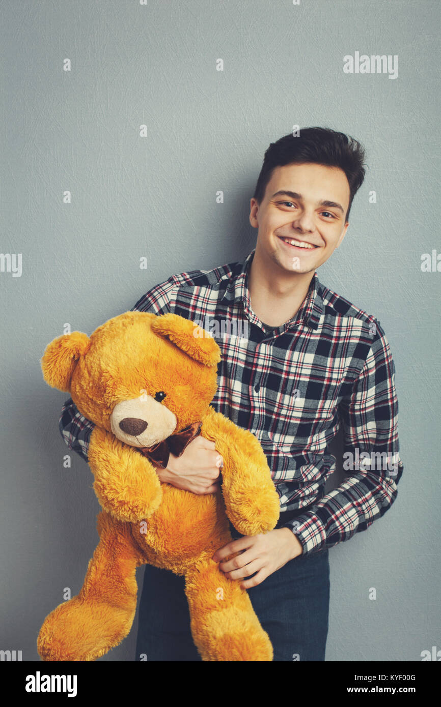 Man holds teddy bear on a light blue background, copy space. Pretty toy concept. Man hugs cute toy bear tight. Guy with happy face plays with red soft toy. Vintage filter Stock Photo